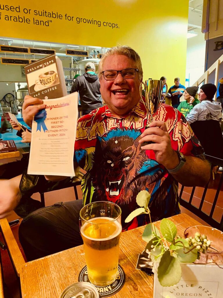 The author, Pat Luther, wearing a colorful shirt with a werewolf across the front. He (Pat, not the werewolf) is holding his novel, Yellow Tape and Coffee, in one hand, along with his certificate and a plastic trophy in the other.