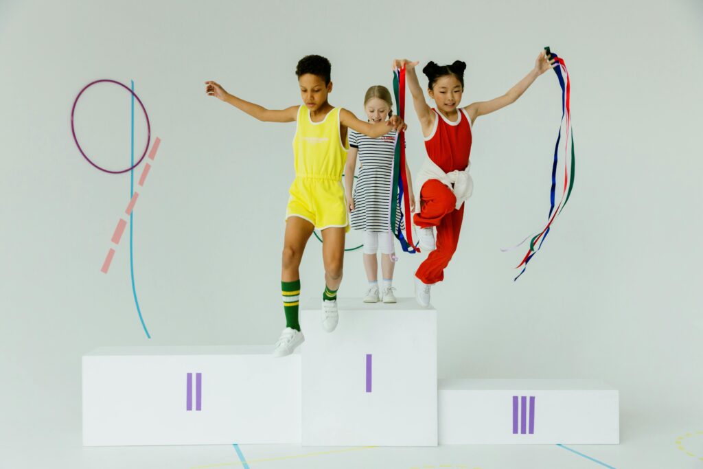 Three kids having fun jumping off a podium with colorful ribbons indoors.